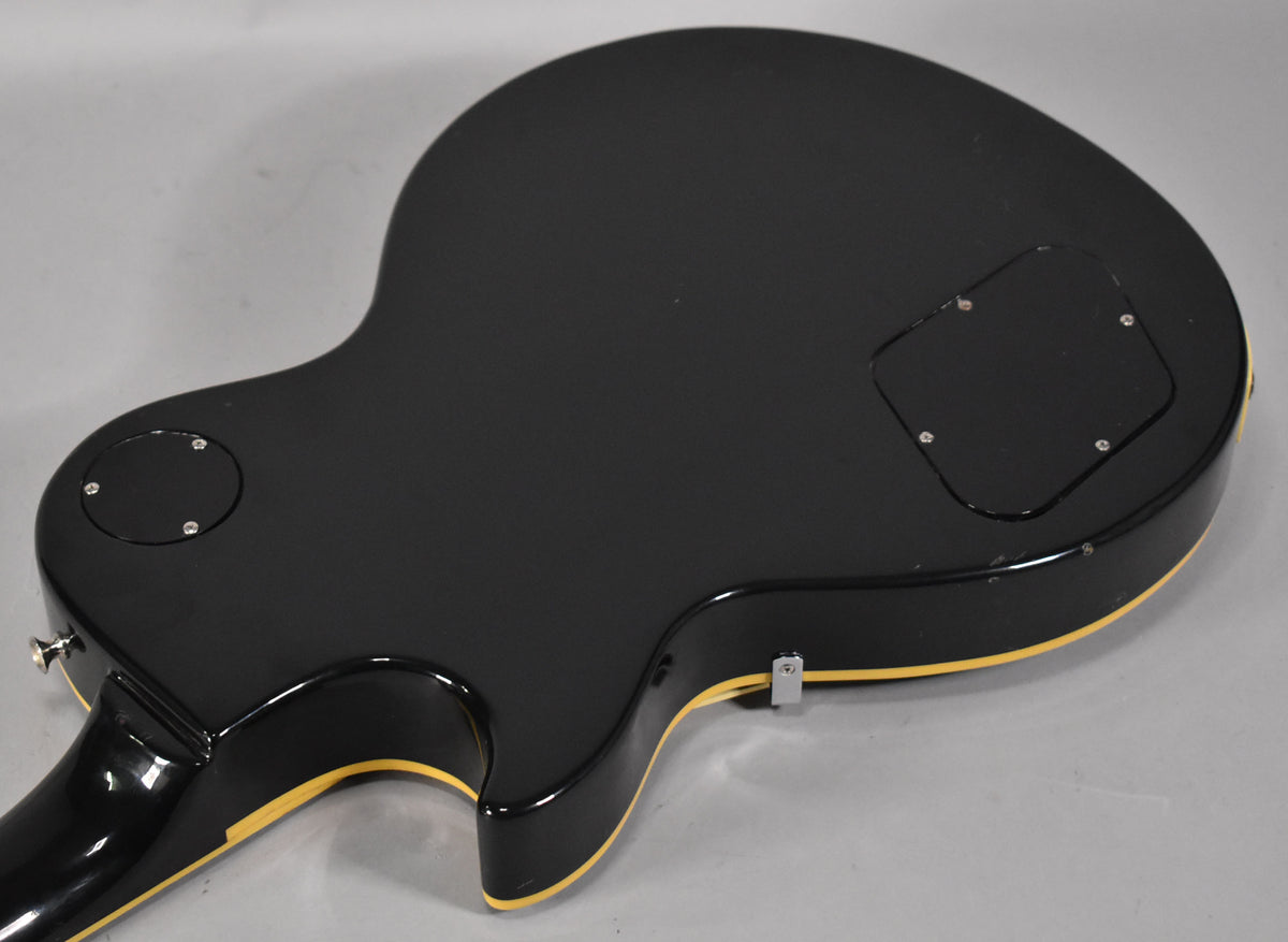 The black guitar's back, angled from the lower bout, shows a control cavity cover, a circular access plate, and cream binding.