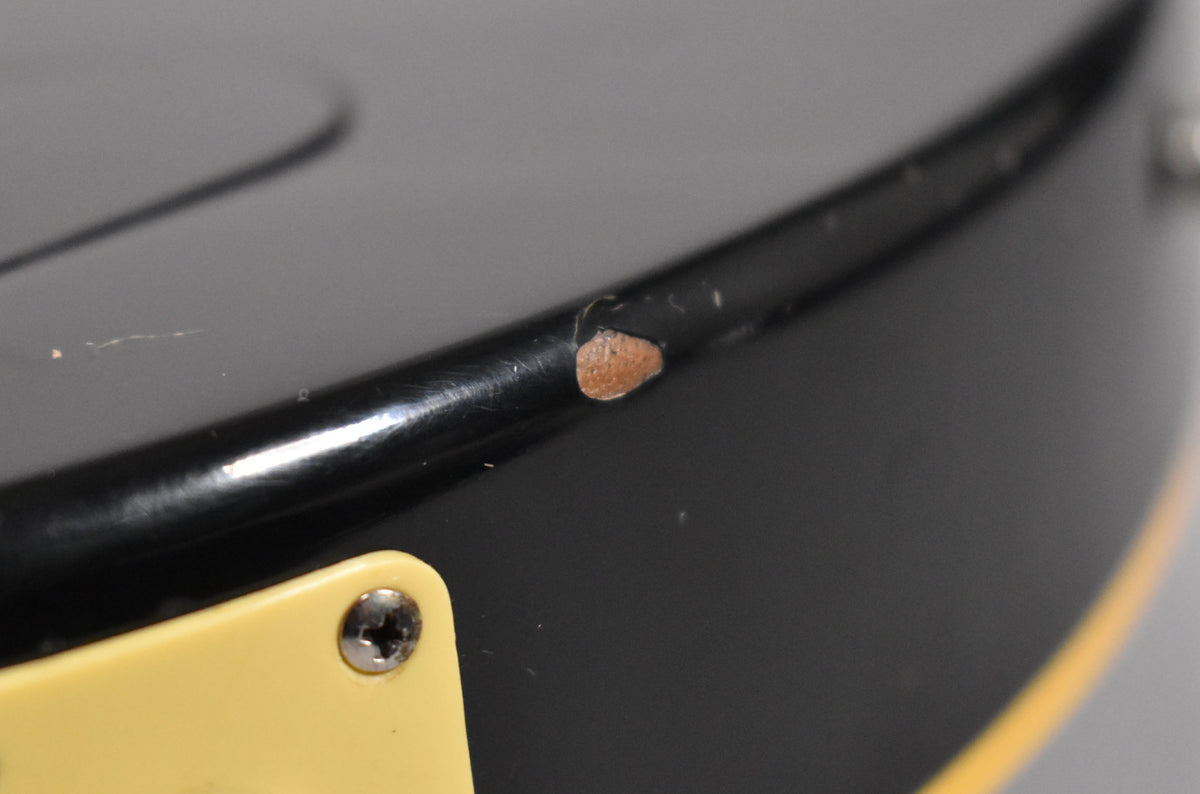 Close-up of the guitar's black body edge, showing a large chip in the finish revealing the wood beneath, and a cream pickguard corner.