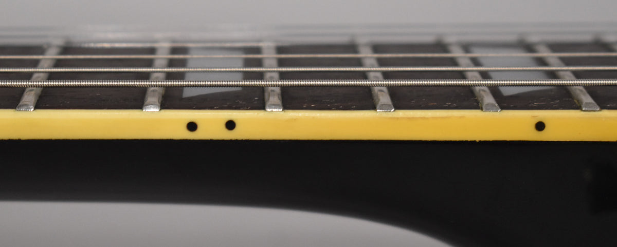 Close-up of the fretboard's side, showing cream binding with black dot markers, dark wood, frets, and strings, with some wear.