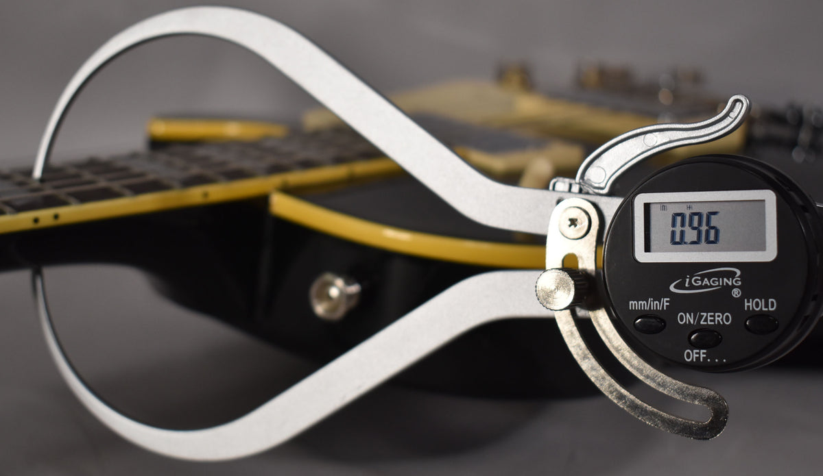 A digital caliper measures the black guitar's fretboard, showing 0.96mm, with yellow binding visible along the body edge.