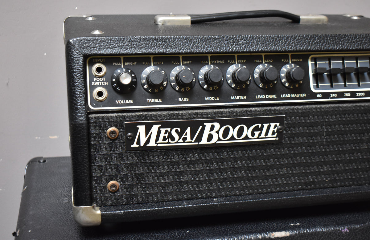 The black textured amplifier head is angled slightly, showing its front panel with many black knobs, an equalizer, and a metal logo.