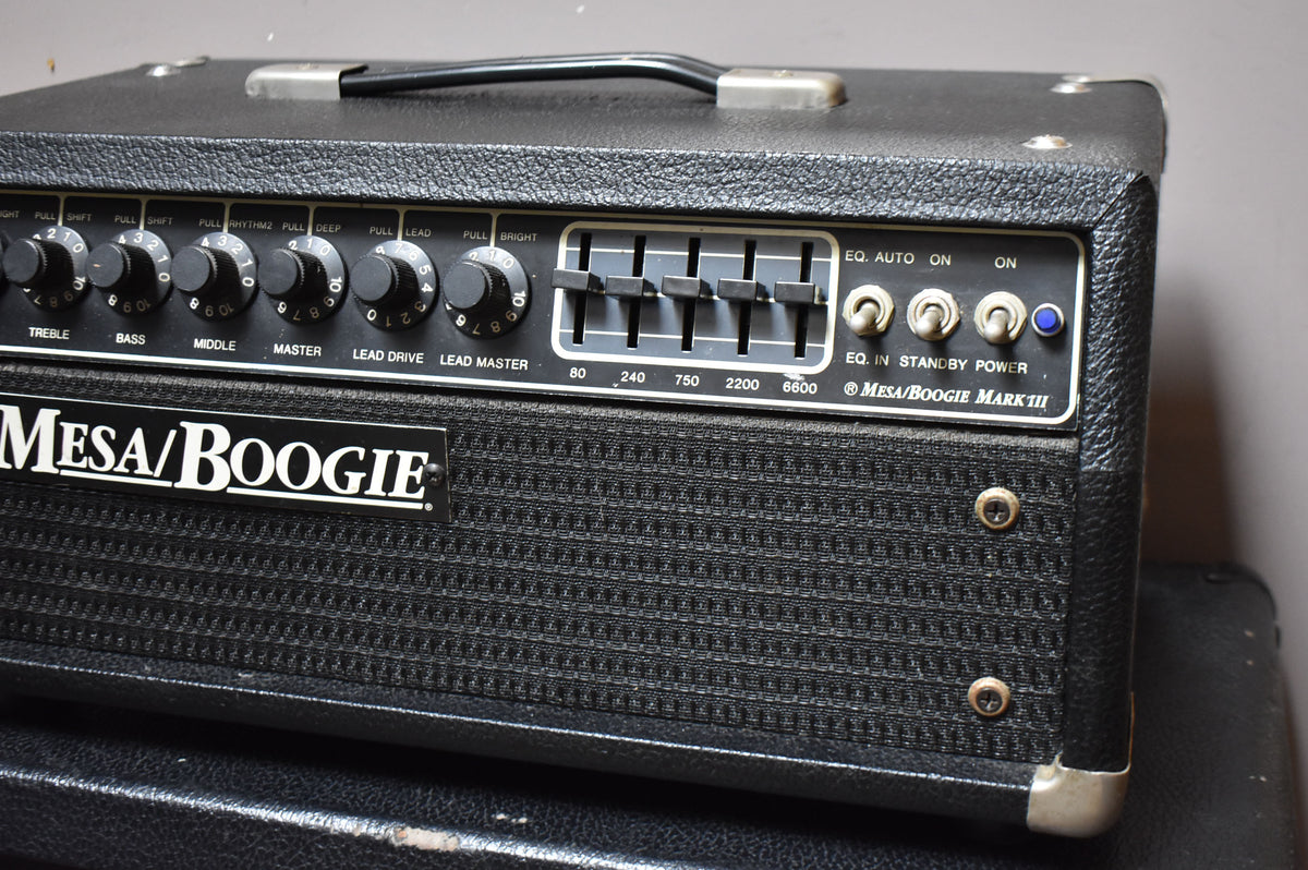 A black textured amplifier head, angled from the right, showing the front panel with numerous black knobs, a 5-band EQ, and a blue power light.