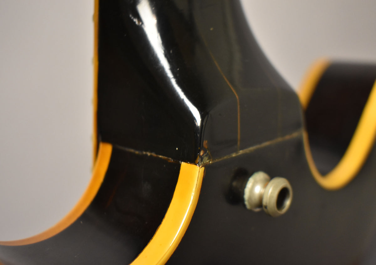 Close-up of a guitar's black body and neck joint, showing cream binding, finish cracks, and a chrome strap button.