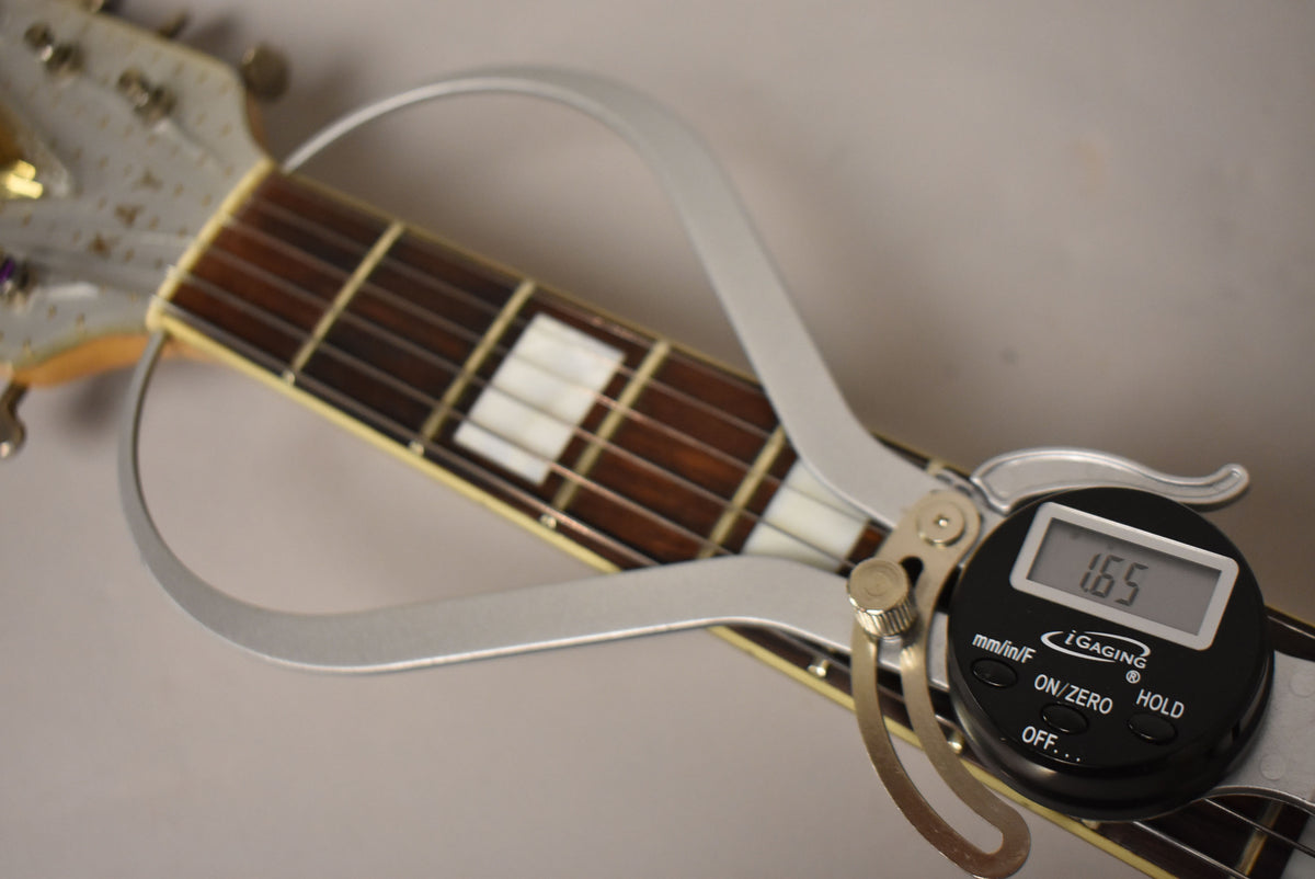 A digital caliper measures the guitar's nut width at 1.65 inches, showing the dark fretboard with rectangular white inlays.