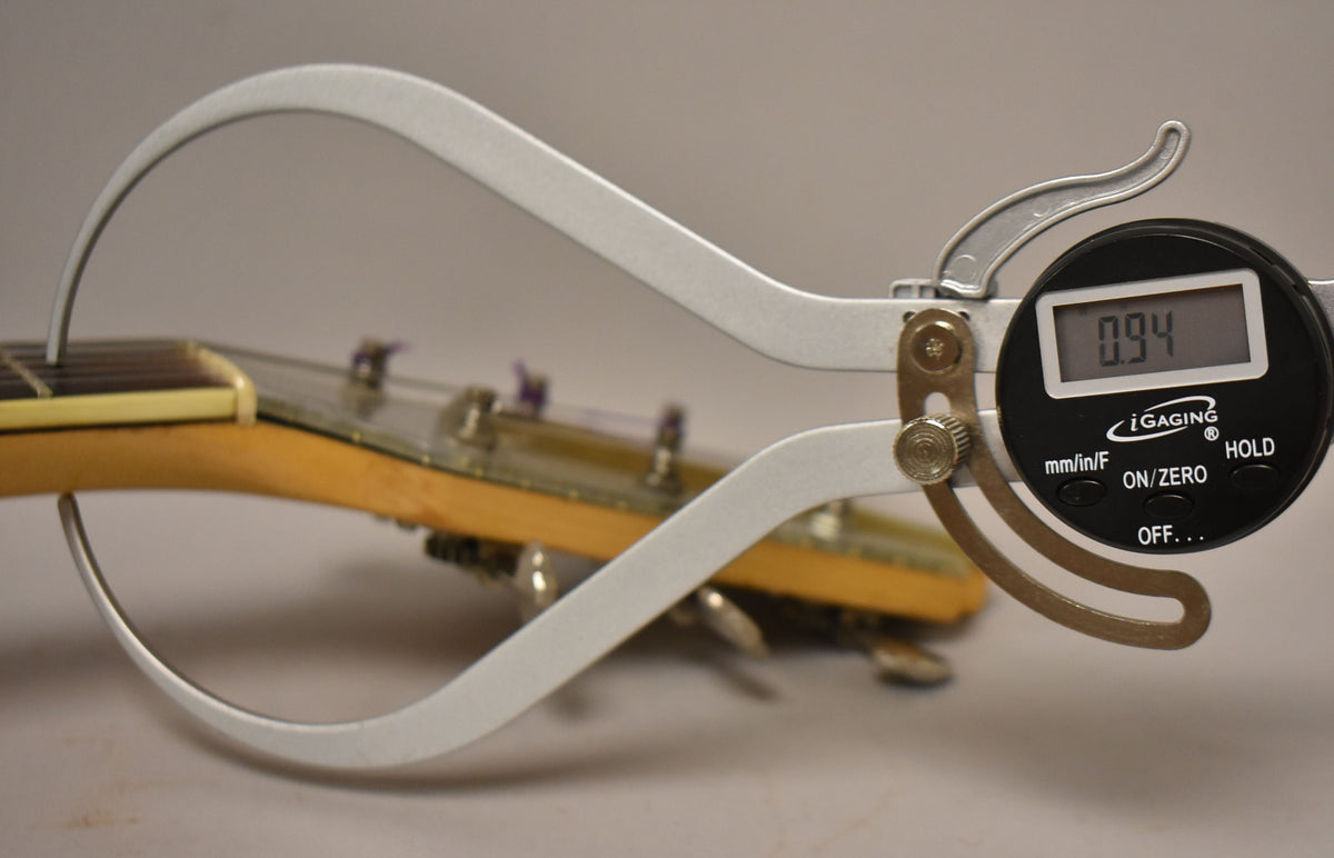 A digital caliper measures the guitar's nut width at 0.94, with the natural finish neck and headstock visible behind it.