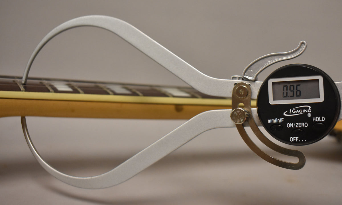 A digital caliper measures the string height at the 12th fret, showing "0.96" on its screen, against the guitar's natural finish neck.