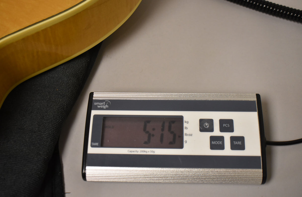 The natural finish guitar body rests on a black case, with a digital scale displaying "5.15" lbs in the foreground.