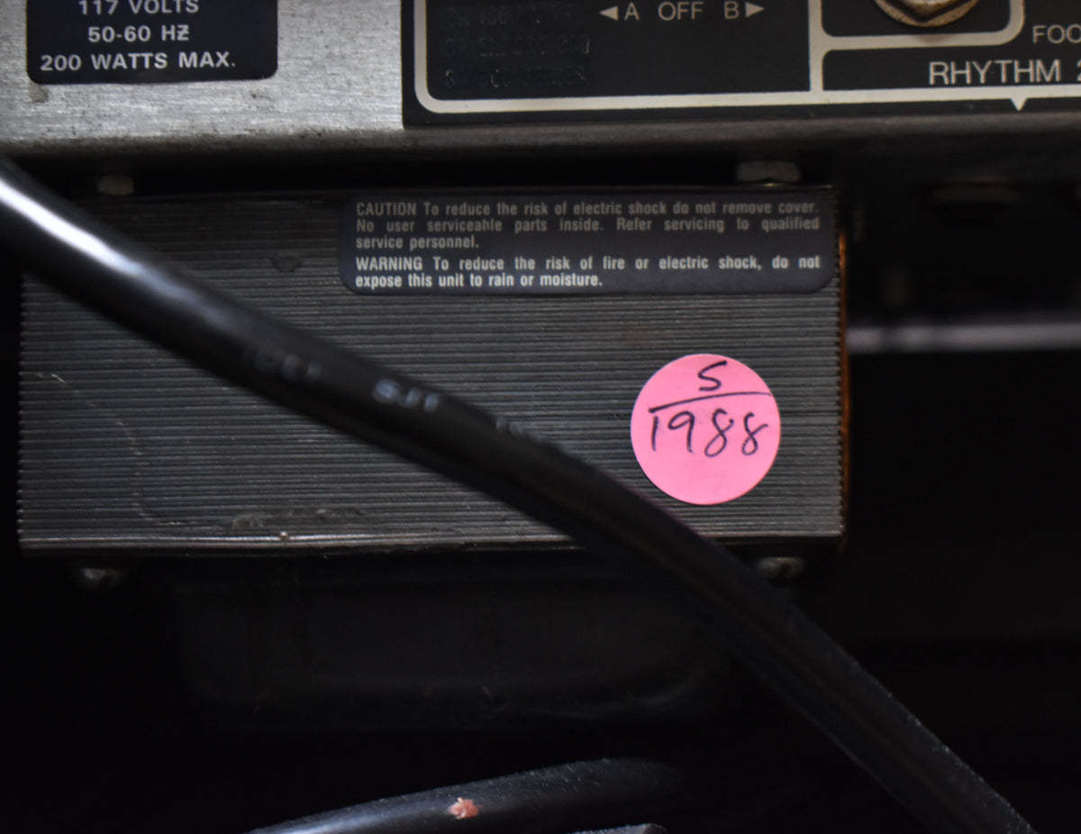 Close-up of the amp's rear, showing a black ribbed metal panel with warning labels and a pink "1988" sticker.