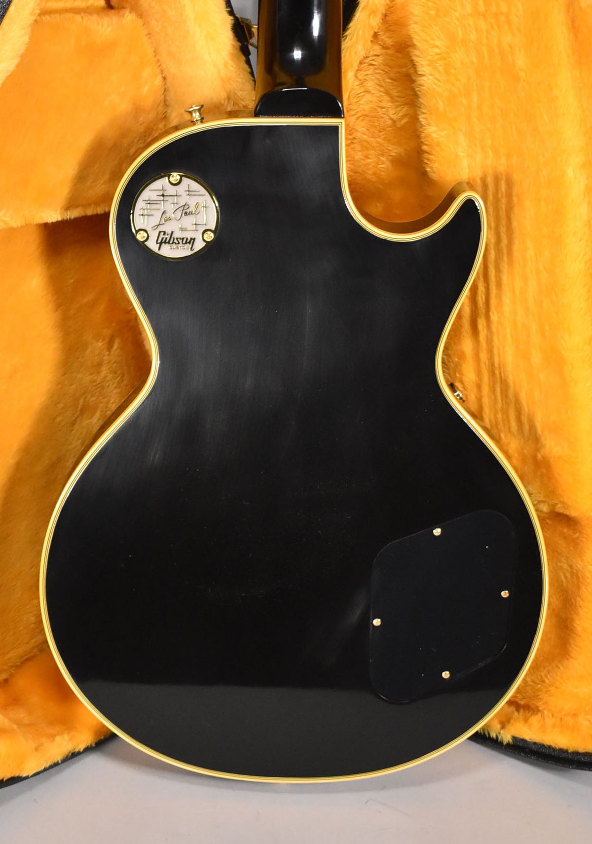The back of a black Les Paul, showing the control cavity cover, neck joint, and a circular backplate with gold screws.