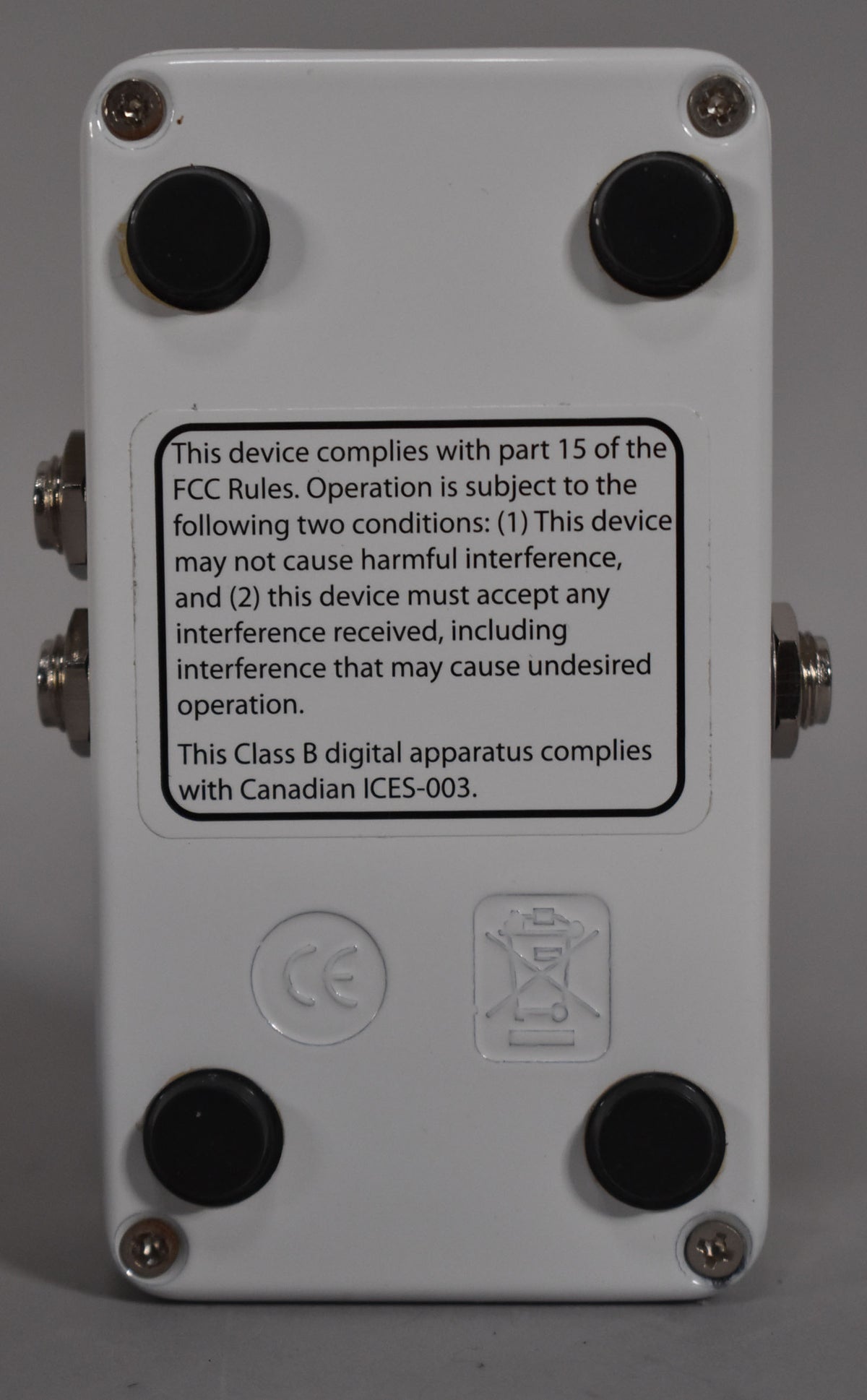 The white pedal's bottom is shown, with four black rubber feet, four silver screws, and a large black and white compliance sticker.