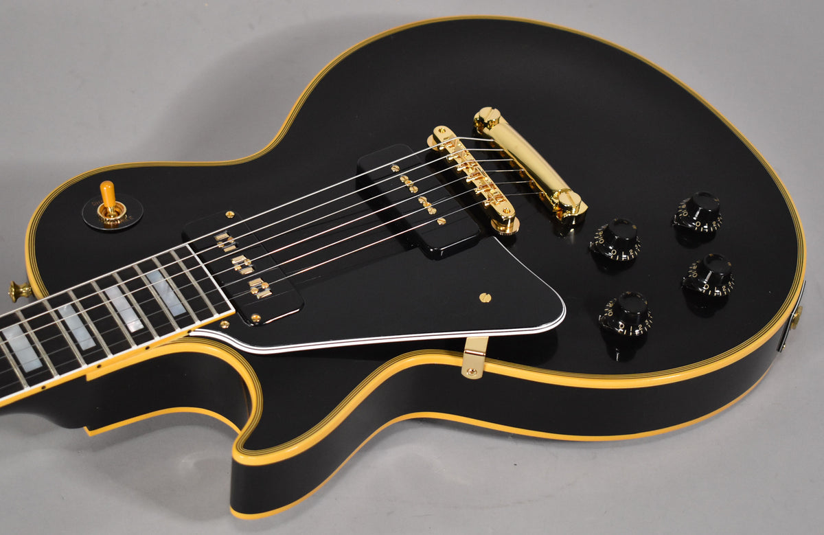 Close-up of a black Les Paul's body, showing gold hardware, black P90 pickups, a black pickguard, and four black control knobs.