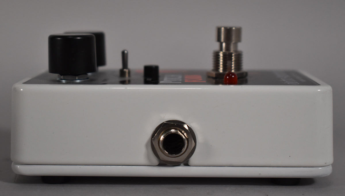 Side view of the white pedal, showing a black knob, a toggle switch, a red LED, a footswitch, and a 1/4-inch input jack.