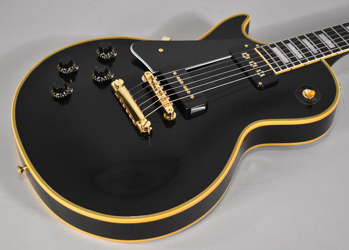 Upper body of a glossy black Les Paul, showing gold hardware, cream binding, four black speed knobs, and block inlays.