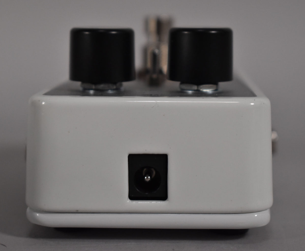 The white pedal is viewed from the front, showing the power input jack and two black control knobs on top.