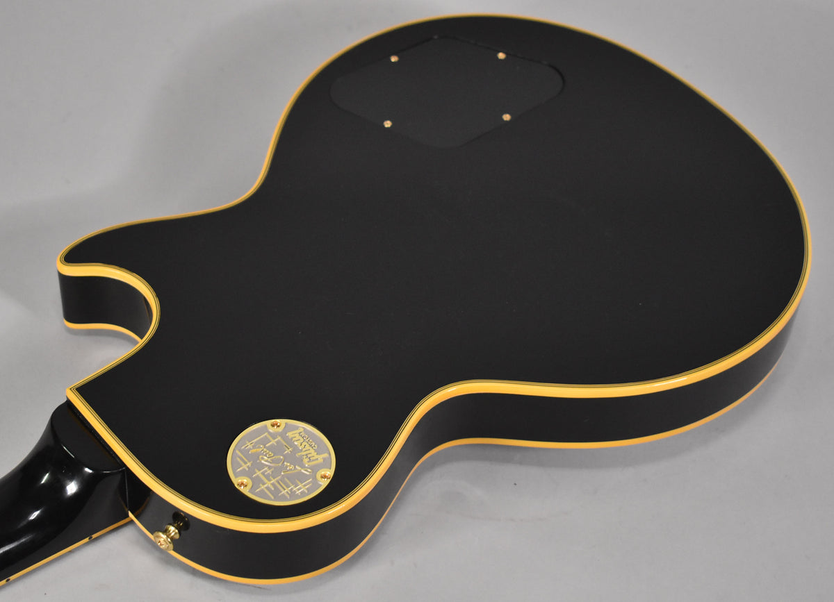 The guitar's matte black back with cream binding, showing the control cavity cover and a gold medallion near the neck heel.