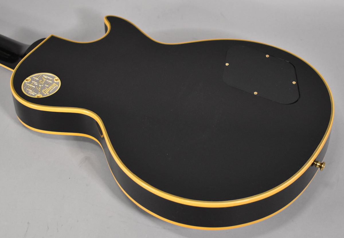 The back of the matte black Les Paul body is angled from the neck joint, showing cream binding and a gold-plated medallion.