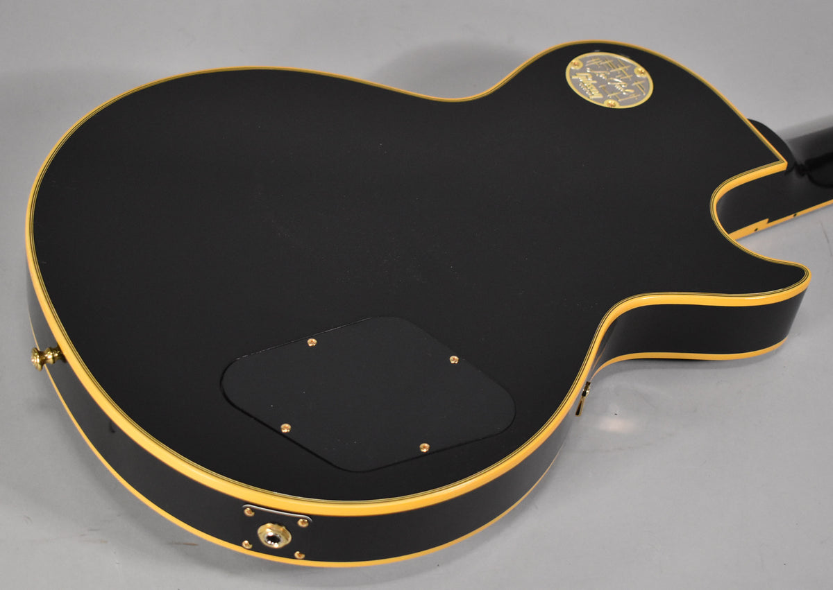 The back of a black Les Paul, showing the control cavity cover, output jack, and a gold medallion on the upper bout.
