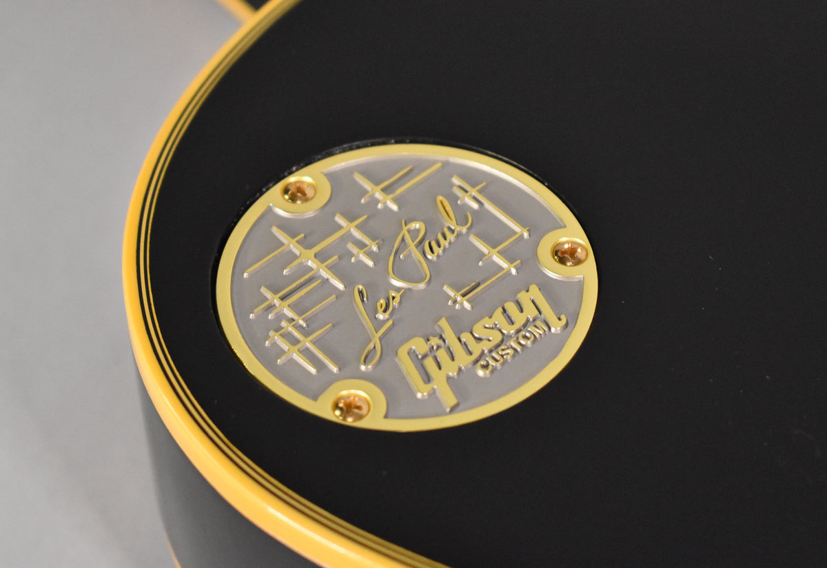 Close-up of the guitar's black body, showing a round silver and gold medallion with four gold screws, and cream binding with a thin black stripe.