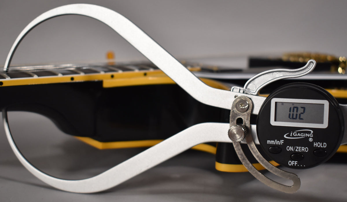A digital caliper measures the guitar's ebony fretboard, showing a 1.02mm reading, with yellow binding visible along the body.