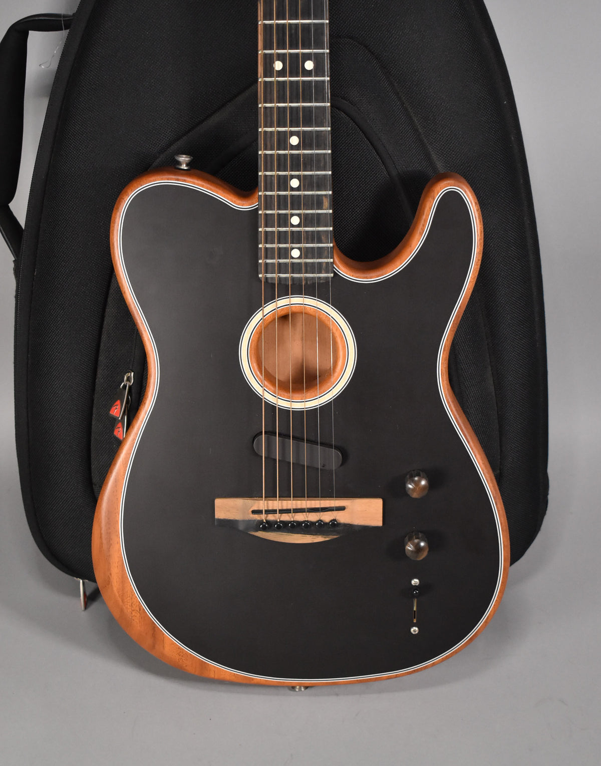 A black matte acoustic guitar with a natural wood binding, soundhole, and bridge, shown upright against a black gig bag.
