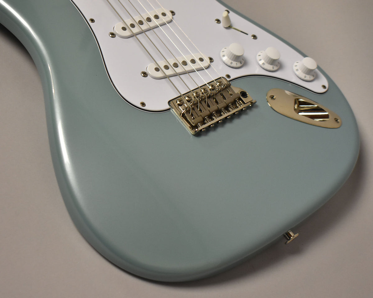Close-up of a glossy polar blue electric guitar body, showing the white pickguard, three white control knobs, gold bridge, and input jack.