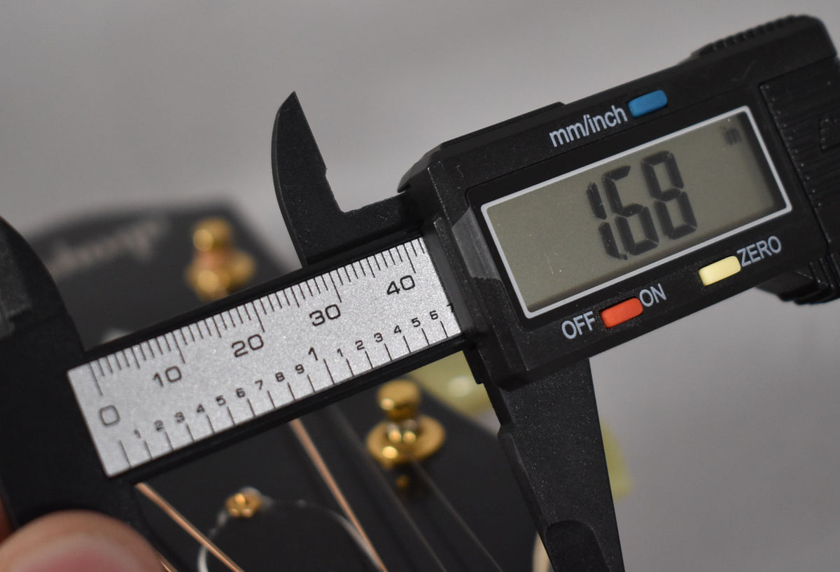 A digital caliper measures the nut width of the guitar's black headstock, showing 1.68 inches, with gold tuning pegs visible.