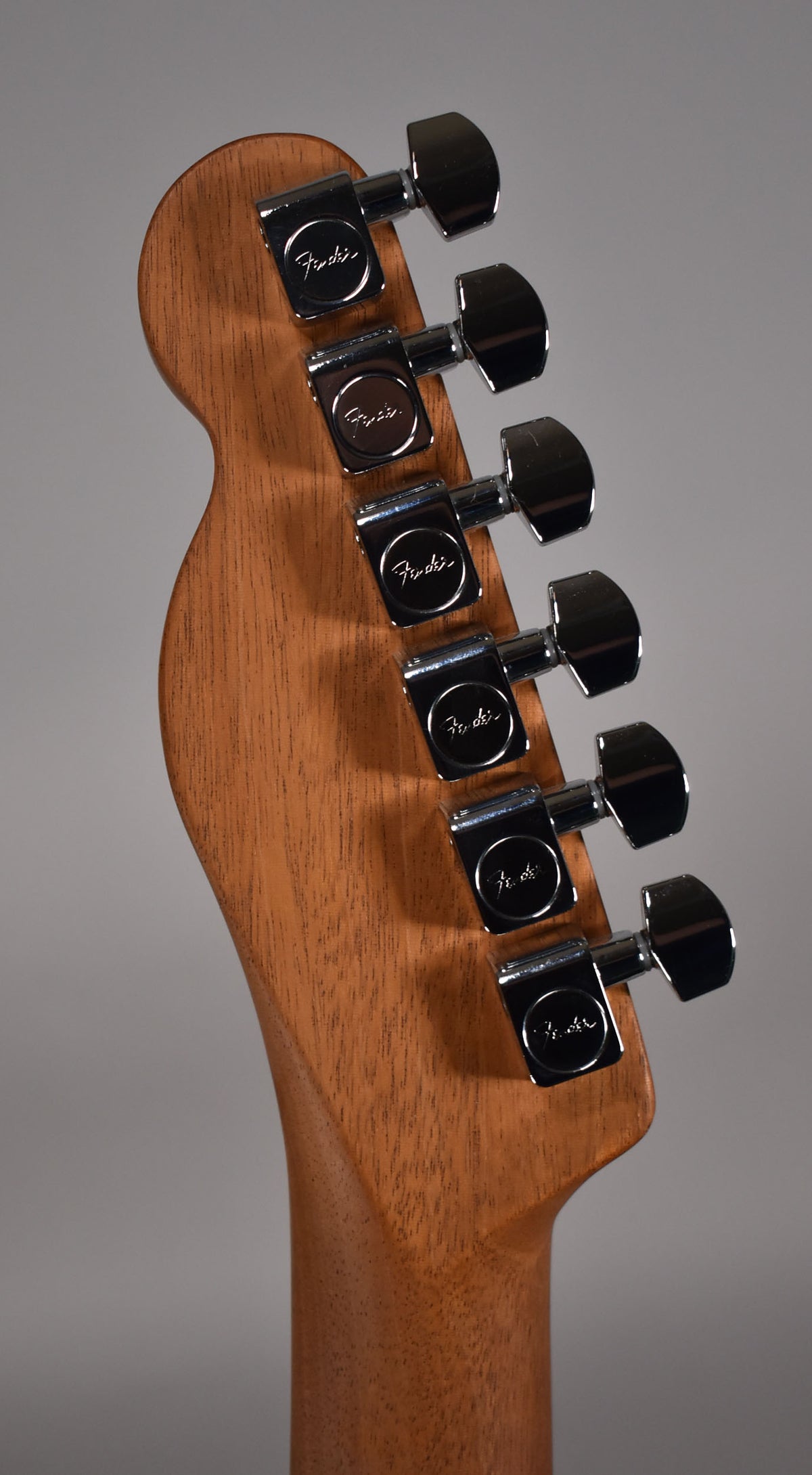 The back of a light brown wooden headstock with six chrome tuners, angled slightly from the left.