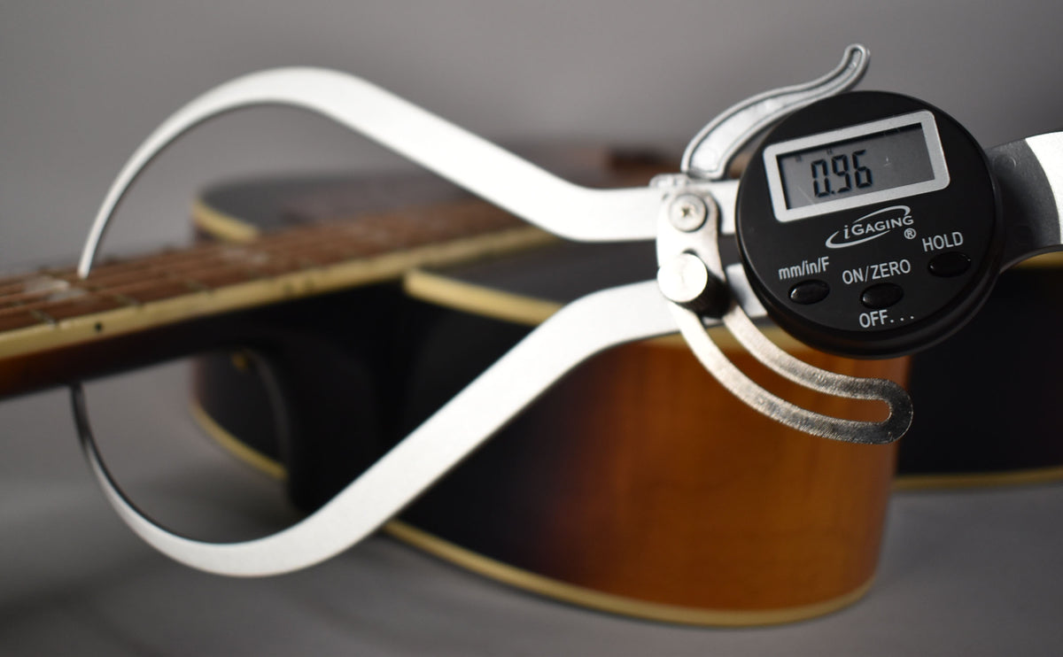 A digital caliper measures the guitar's neck, showing "0.96" on its display, with the sunburst body blurred in the background.