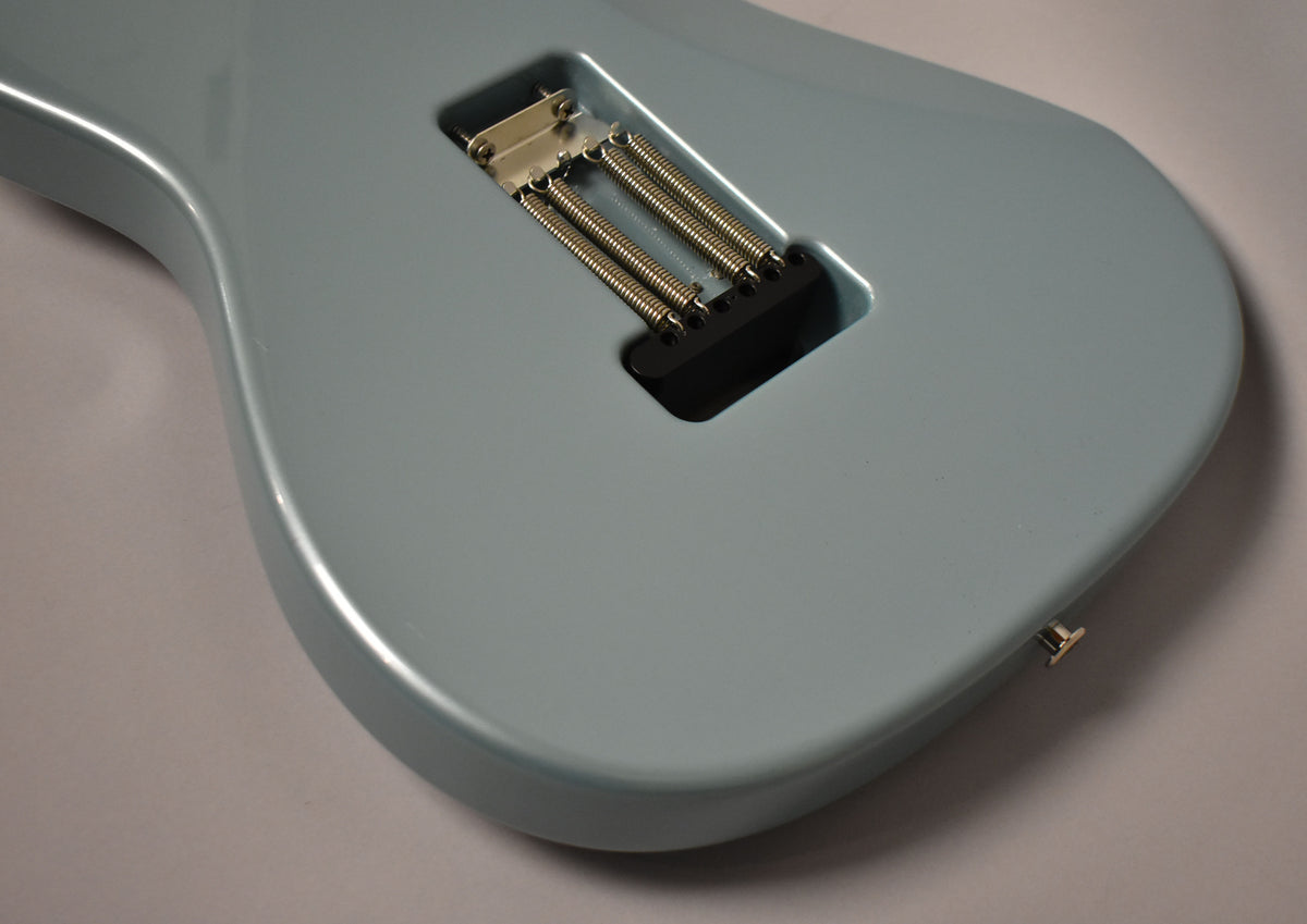 The back of a light blue guitar body, showing the tremolo spring cavity with three springs and a gold strap button.