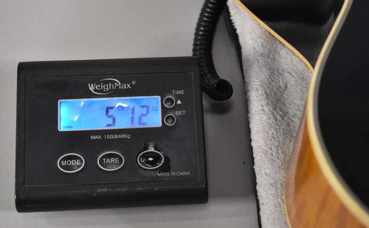 A black digital scale displays "5 LB 12 OZ" next to the lower bout of the guitar, resting on a grey cloth.