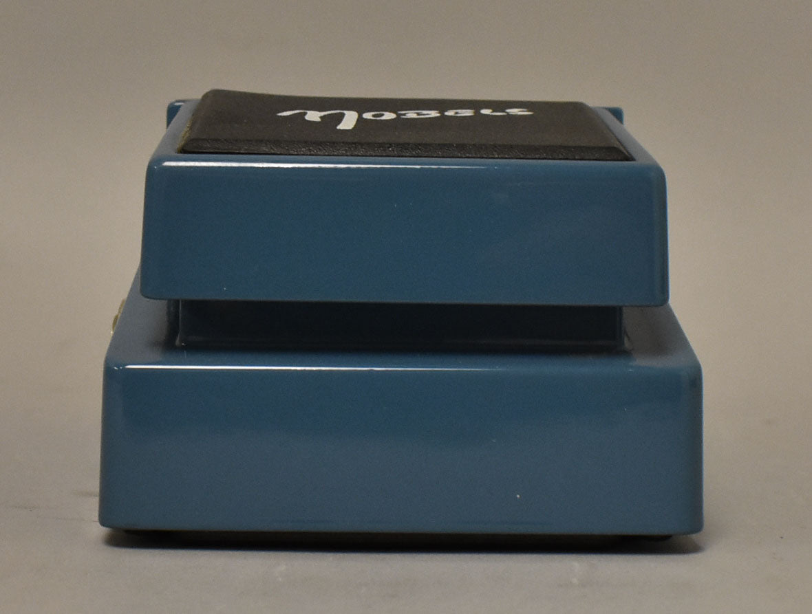 The blue pedal is viewed from the left side, showing the black rubber top with white script and a small gold jack on the lower left.
