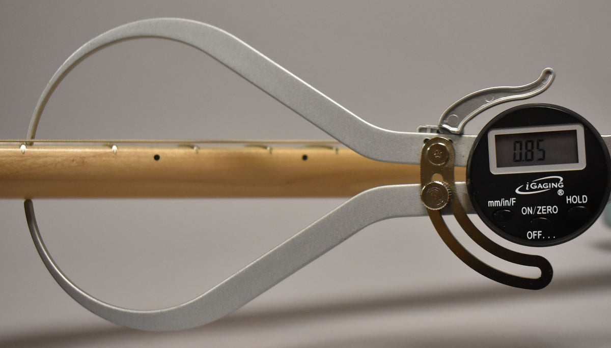 A digital caliper measures the neck thickness of the light-colored maple fretboard, displaying "0.85" on its screen.