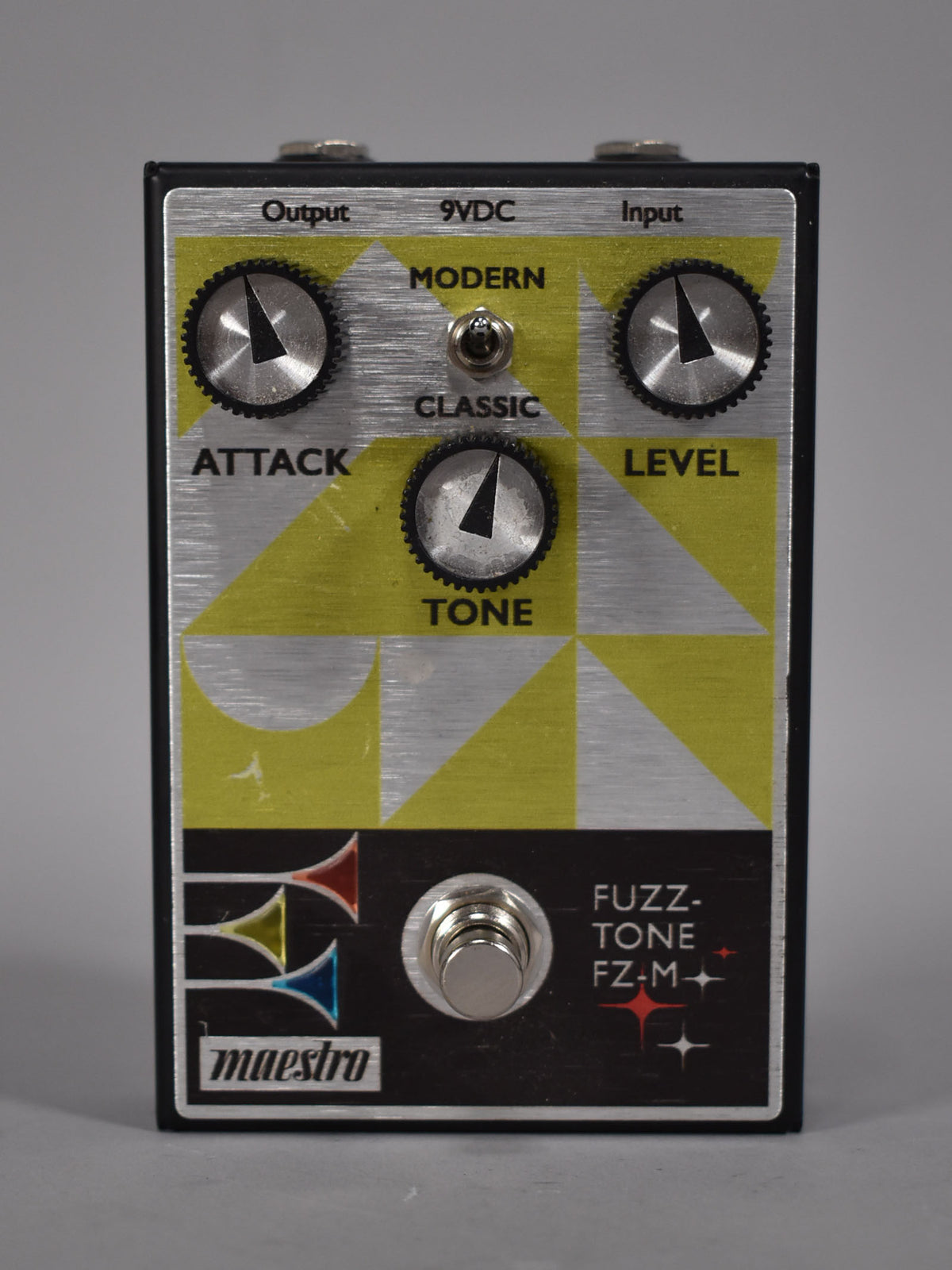 A top-down view of a black pedal with a brushed aluminum faceplate, yellow geometric patterns, three knurled knobs, and a footswitch.