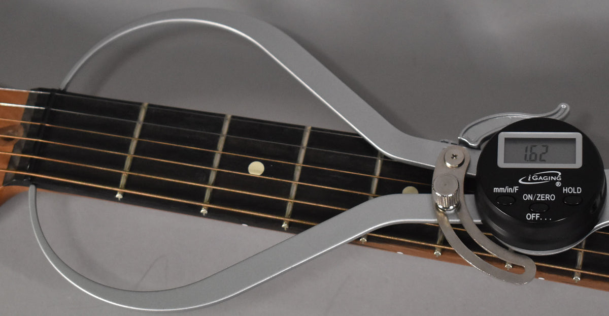Close-up of the guitar's dark fretboard and light strings, with a silver digital caliper measuring the string height at 1.62.