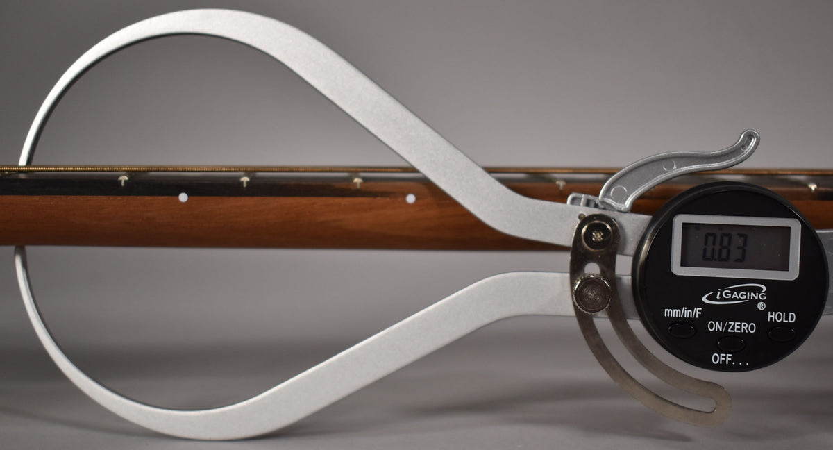 A digital caliper measures the neck relief of the guitar, showing 0.83 on its screen, with the fretboard and strings visible.