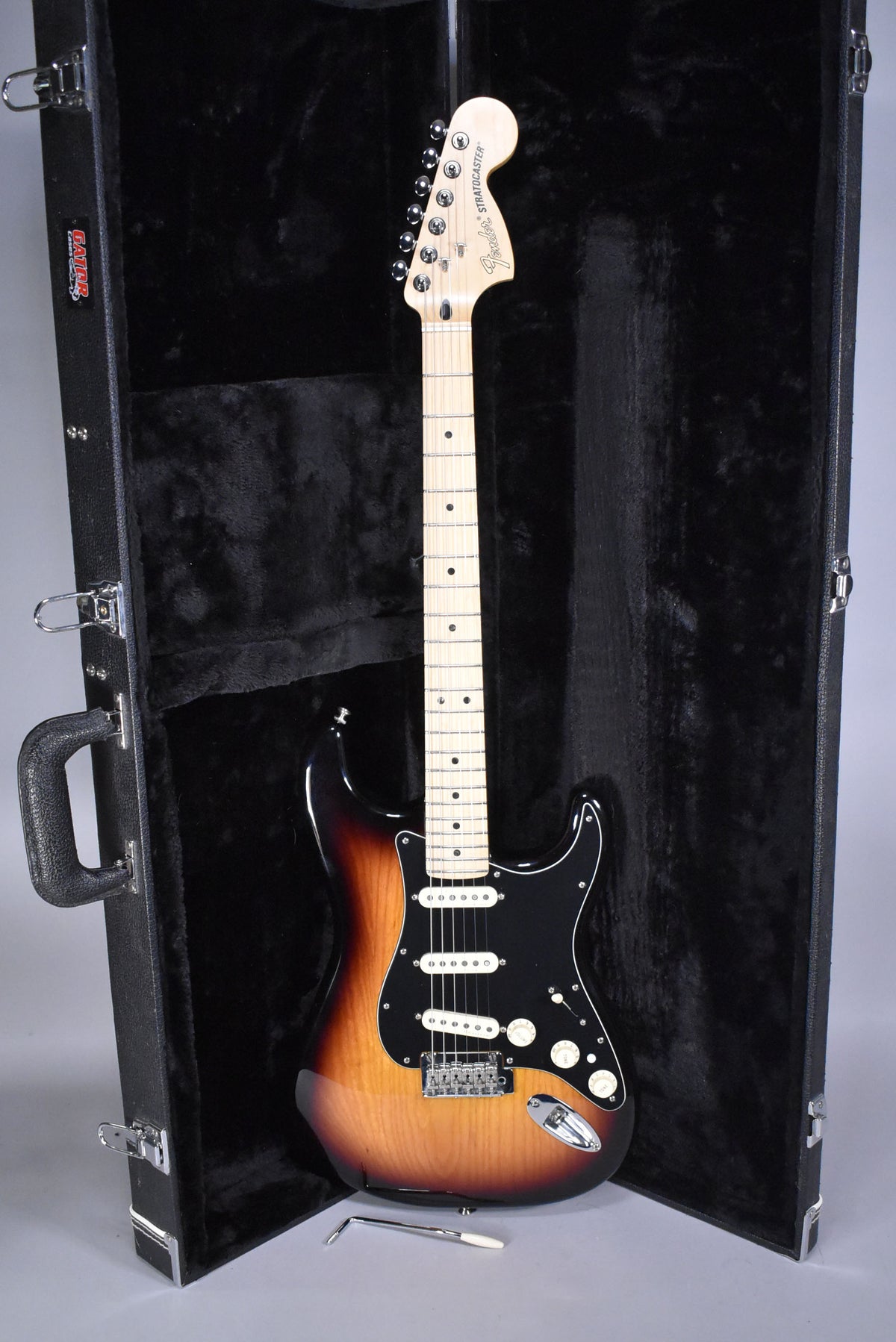 Full-length view of a glossy 3-tone sunburst Stratocaster with a black pickguard, maple fretboard, and white pickups, standing in its open hard case.