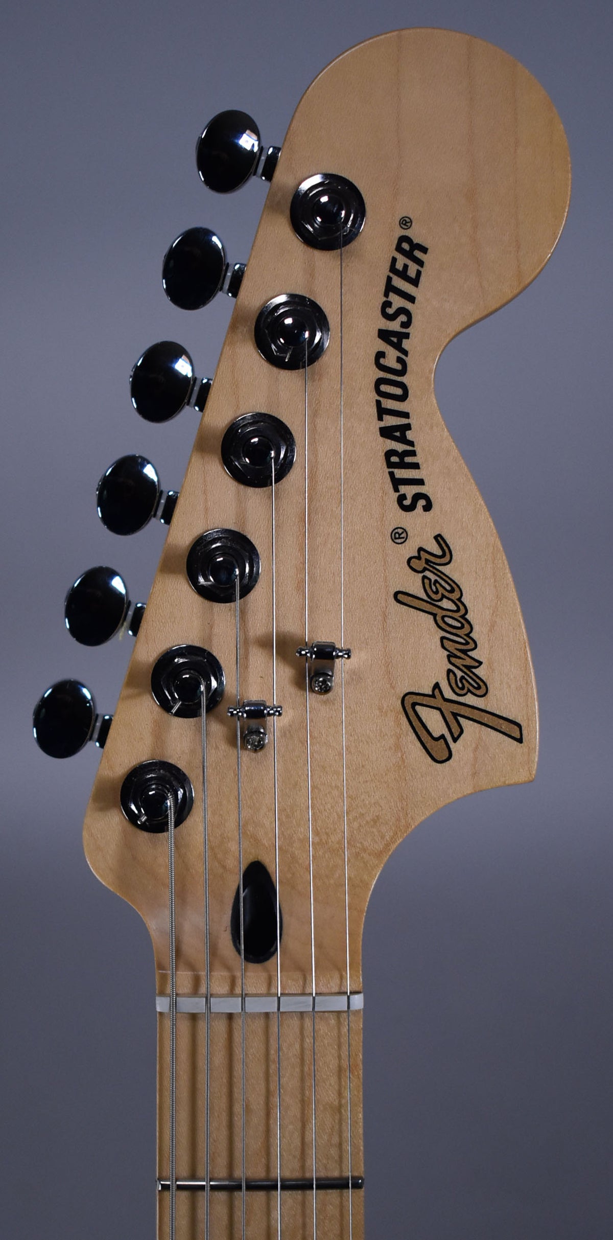 Close-up of the guitar's headstock, angled slightly left, showing six black tuning pegs, two string trees, and a light wood finish.