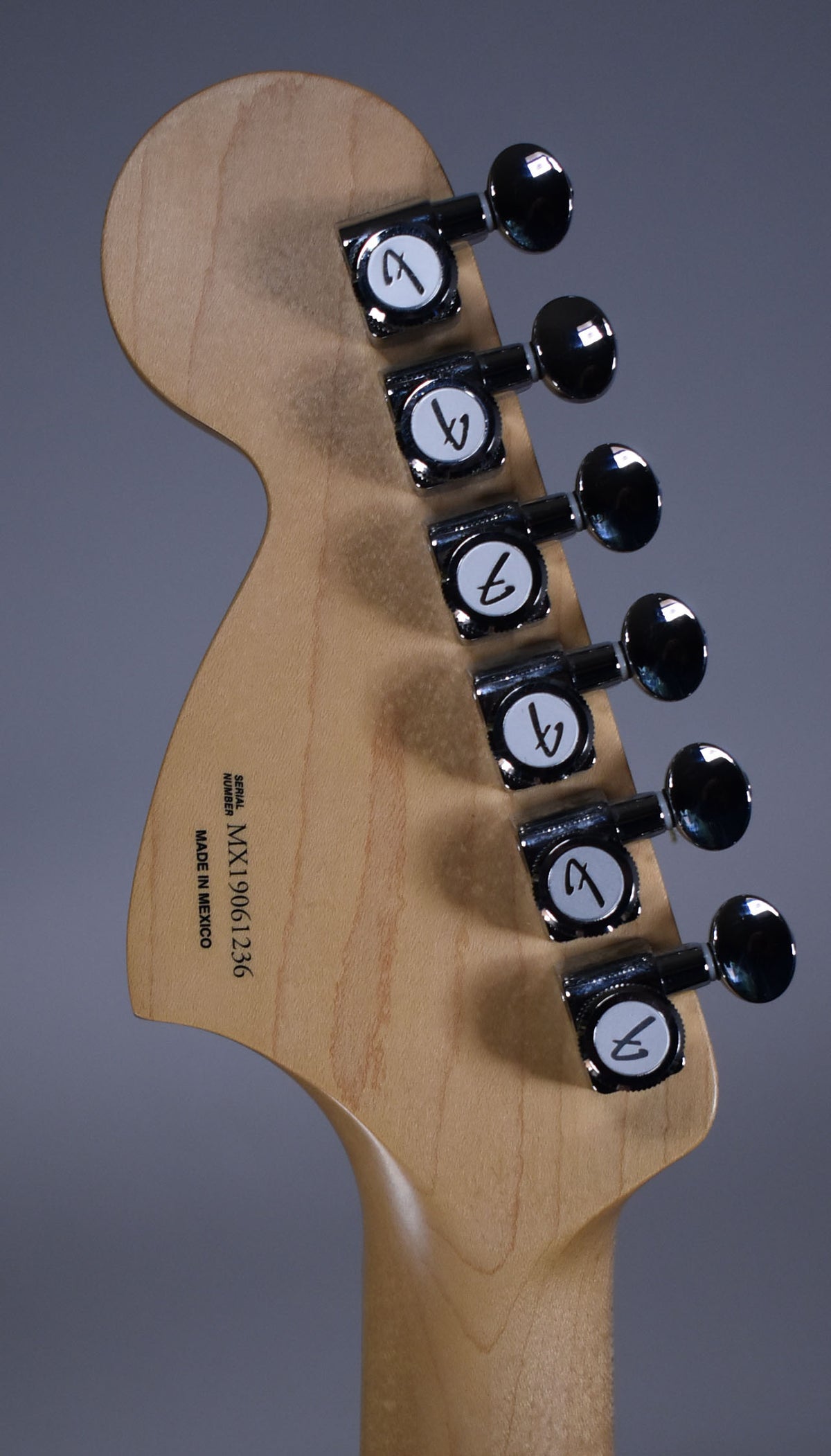 The back of a light maple headstock and neck, angled right, with six black-buttoned chrome tuners in a 6-in-line configuration.