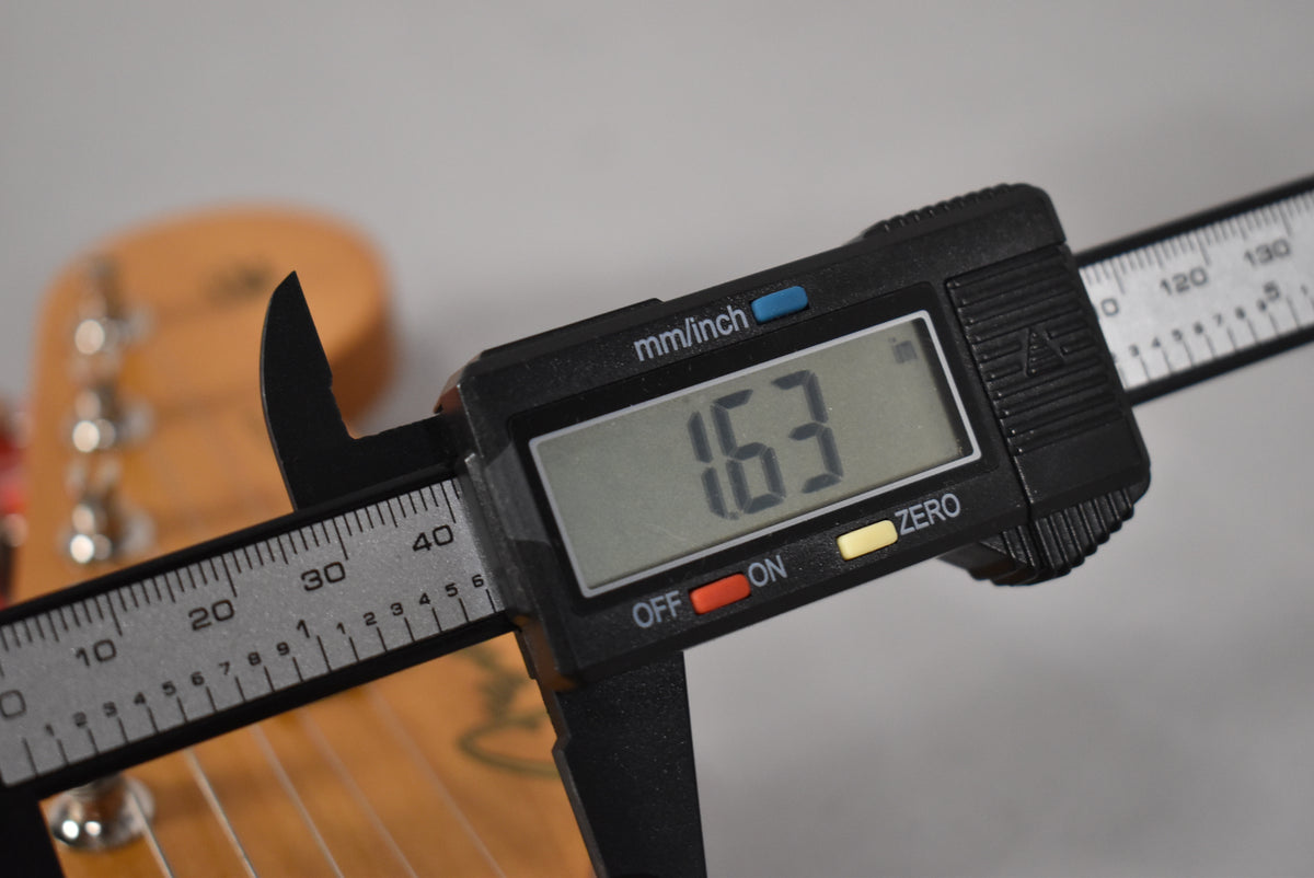 A digital caliper measures the nut width of the guitar neck at 1.63 inches, with tuning pegs visible in the background.
