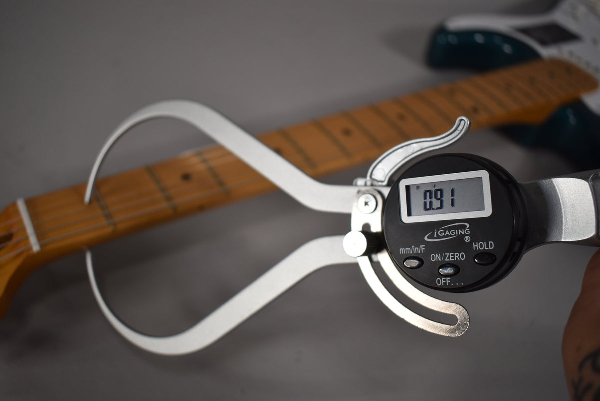 A digital caliper measures the neck thickness of the Stratocaster's maple fretboard, showing "0.91" on its display.