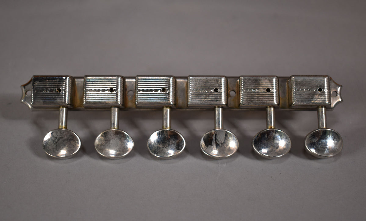 Close-up of six tarnished, silver-colored tuning machine heads with oval buttons, mounted on a metal strip.