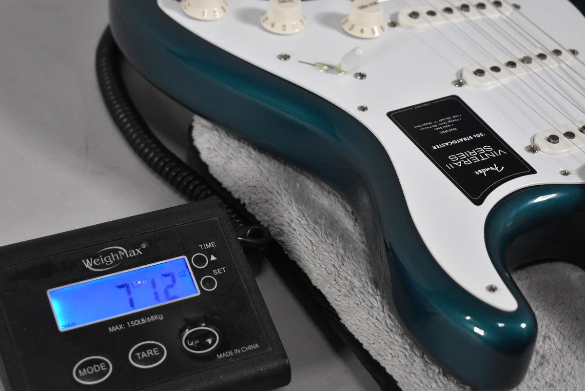 The Ocean Turquoise Stratocaster body, with white pickguard and cream knobs, rests on a scale displaying 7 lbs 12 oz.