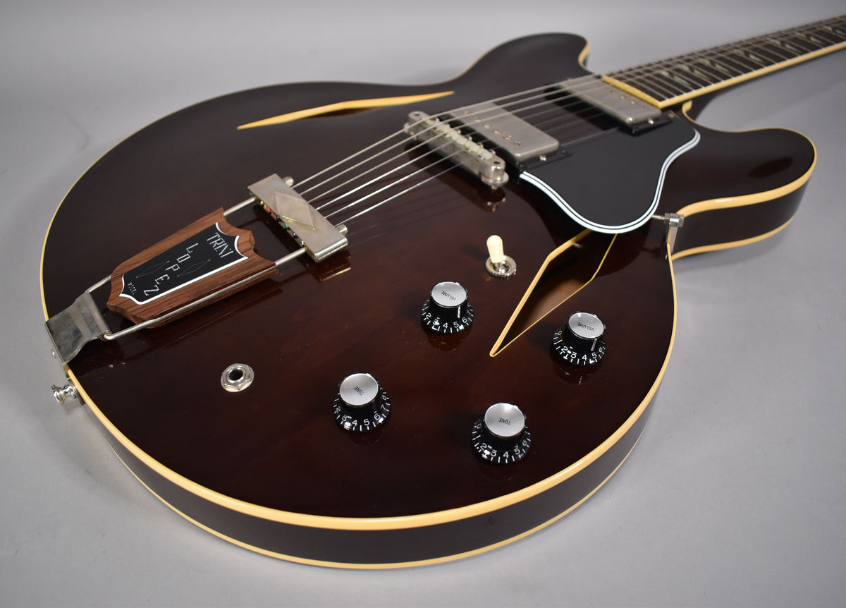 The dark walnut semi-hollow body guitar is angled, showing its four black volume/tone knobs, cream binding, and silver hardware.