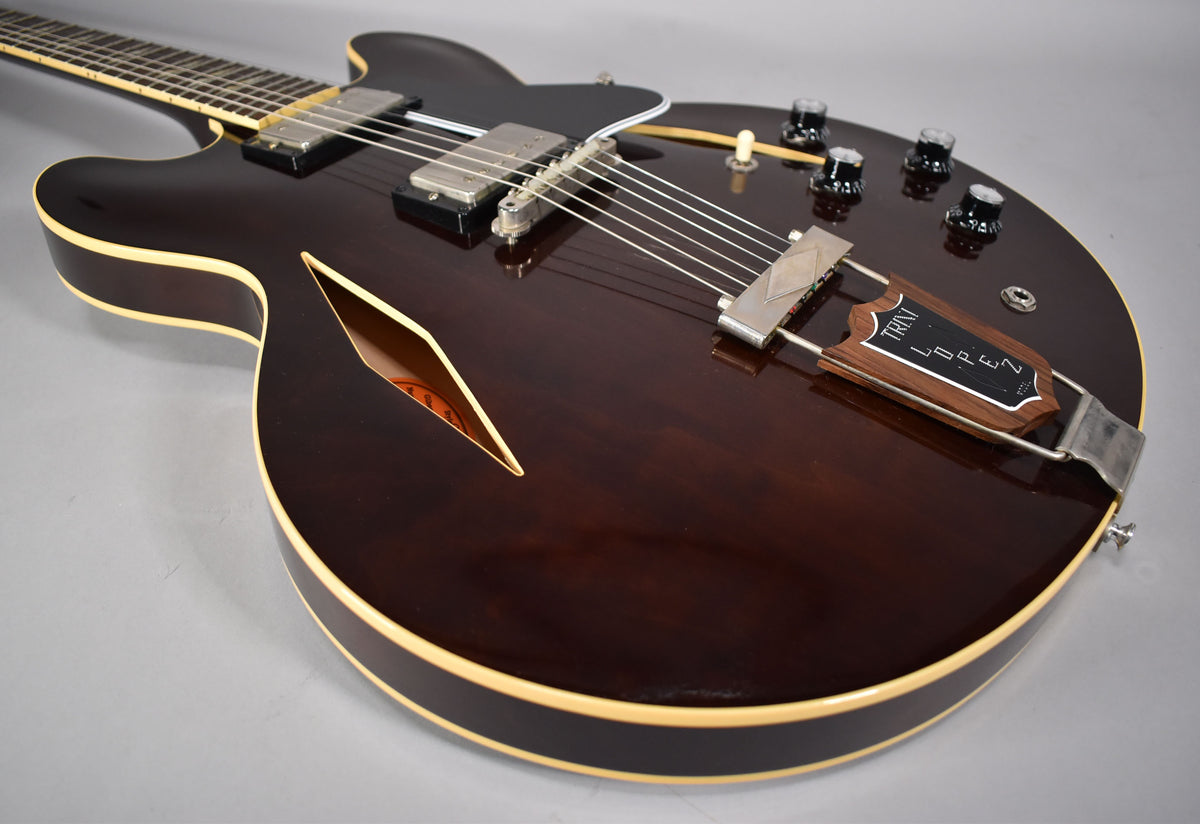 The dark walnut semi-hollow body guitar is angled from the lower bout, showing the f-hole, two humbuckers, and a wooden trapeze tailpiece.