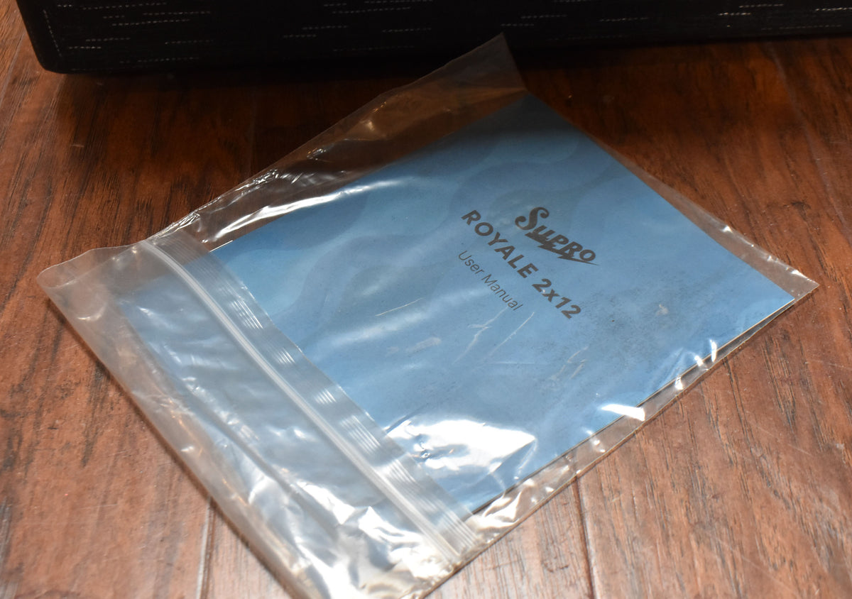 A blue "Royale 2x12 User Manual" is sealed in a clear plastic bag, resting on a dark wooden floor.