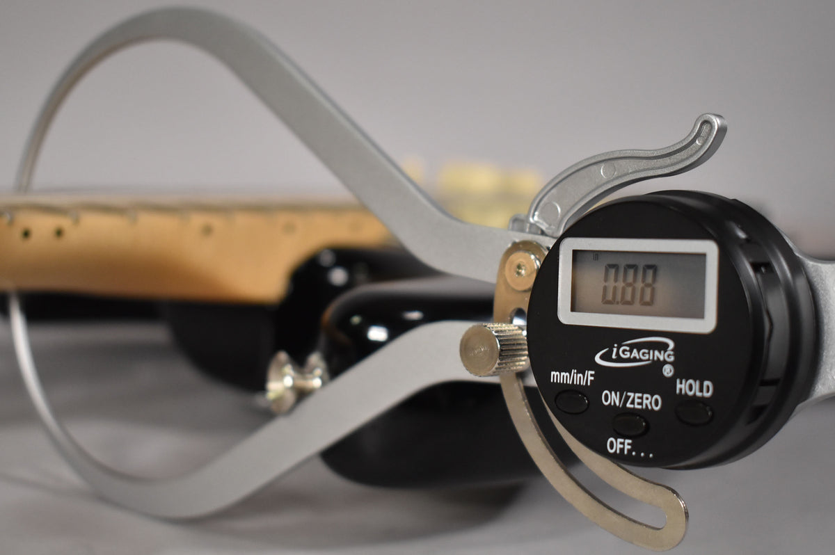 A digital caliper with a reading of "0.00" is shown measuring the neck of a black guitar.