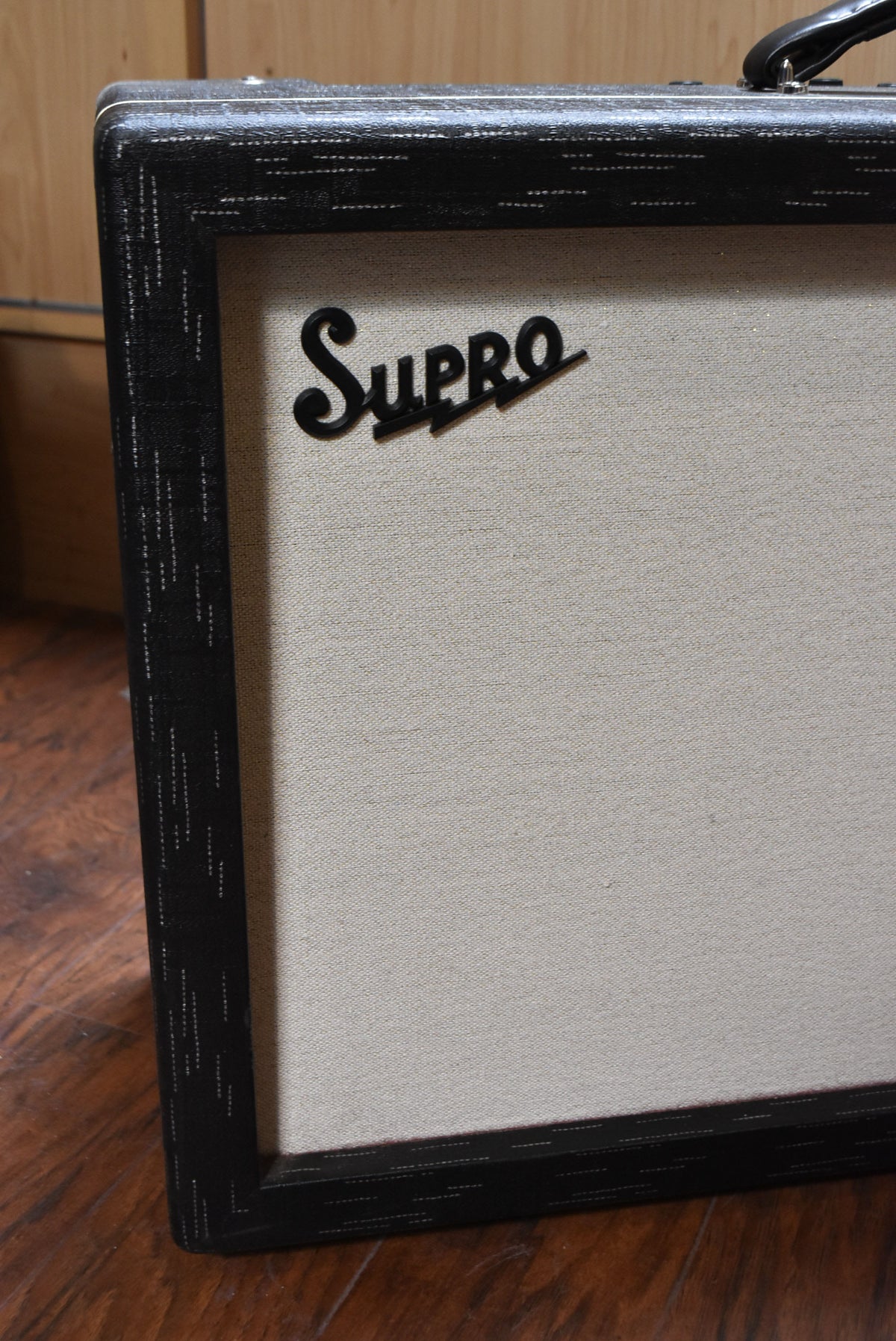 The Supro amp's front is angled, showing its light beige grille cloth and black textured frame with white flecks, plus a black Supro logo.