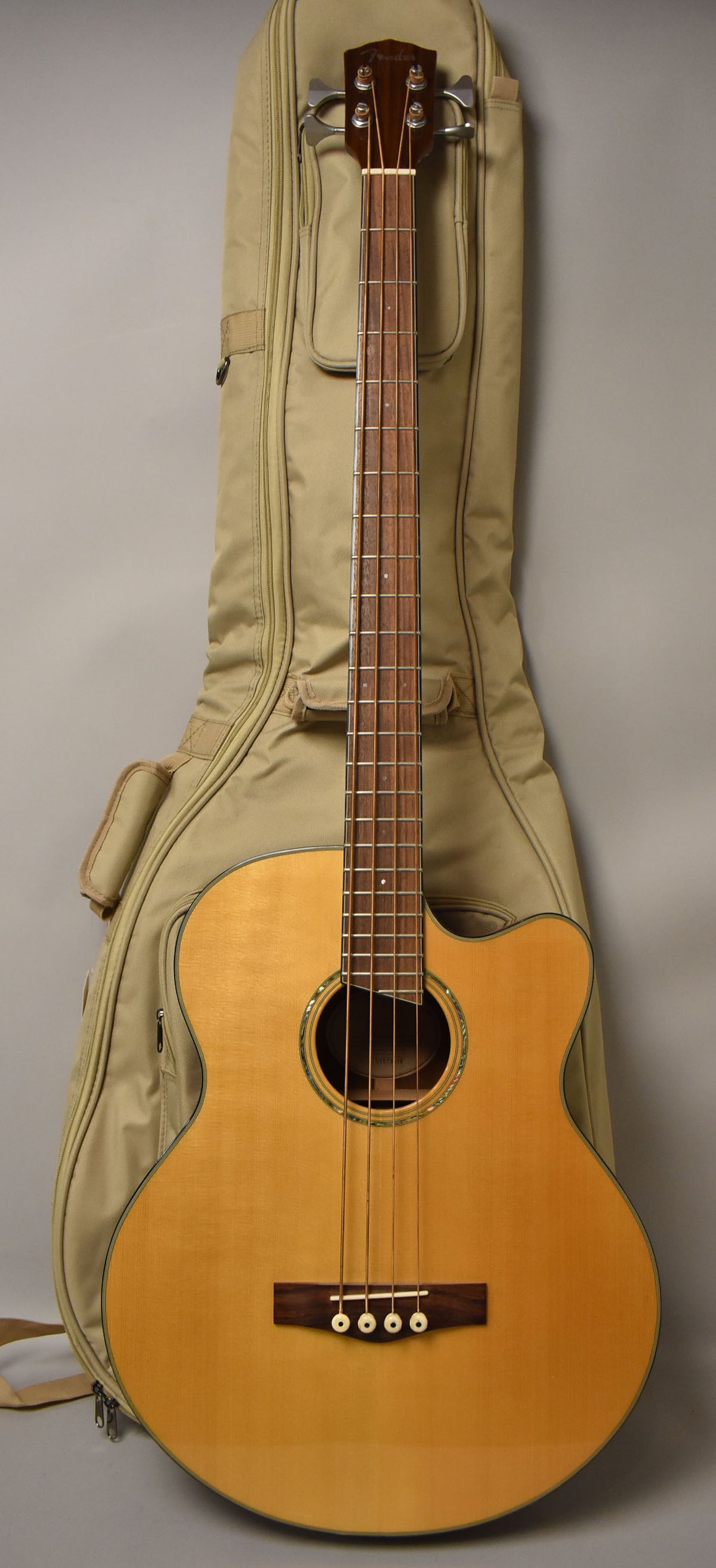 Full-length view of a natural finish acoustic bass with a cutaway body, dark fretboard, and a tan gig bag behind it.