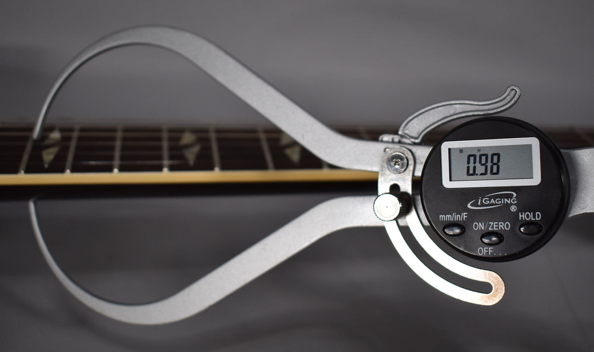 A digital caliper measures the guitar's neck, showing 0.98 on its display, with trapezoid inlays visible on the fretboard.
