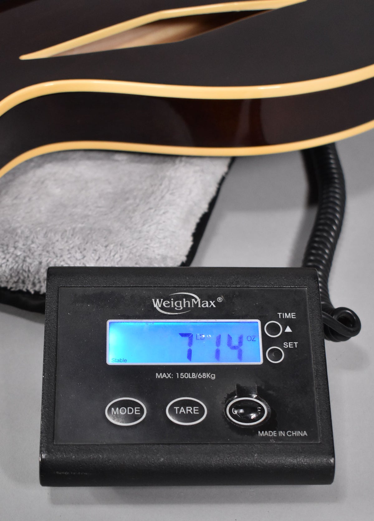 The guitar's dark walnut body with cream binding rests on a scale displaying "7 LB 14 OZ" on a blue backlit screen.